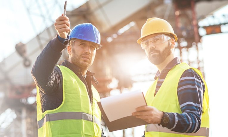 Two engineers with hard hats in a site. One is looking and pointing up a pen and one is looking up holding a clipboard.