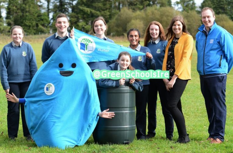 A group of students and their teacher smiling with the green schools blue flag and the mascot of green schools