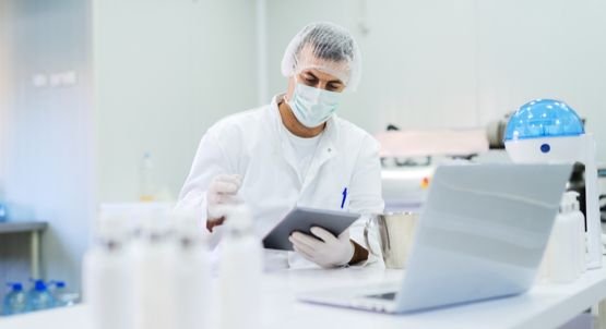 A lab worker wearing a mask and hair net while sitting at a desk with an electronic tablet in hand