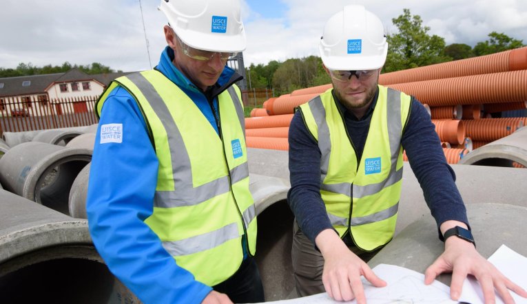 Two Irish Water engineers looking at plans outside storing large concrete sewerage pipes.