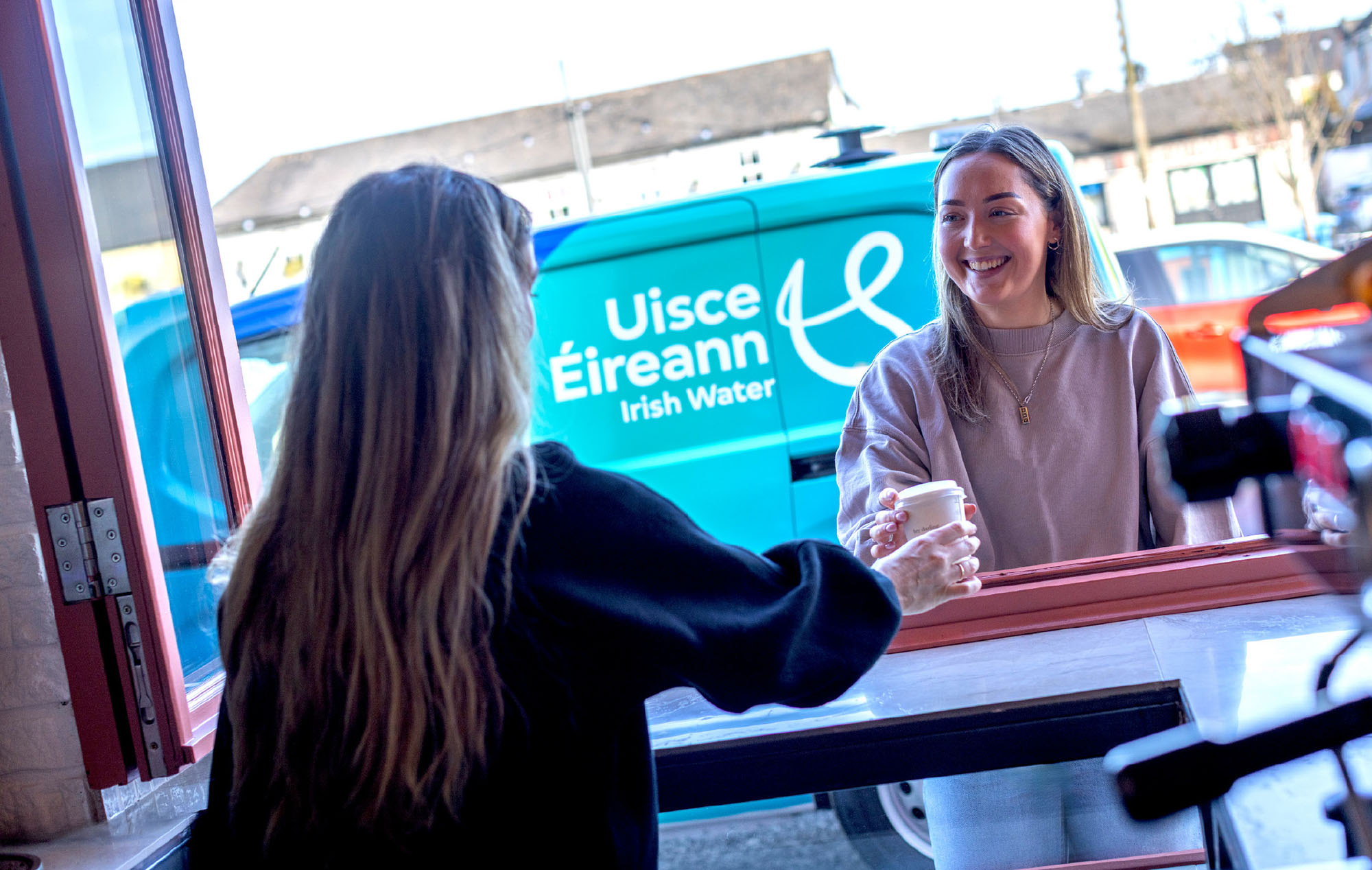 Barista ag freastal ar chustaiméir mná trí haiste i gcaifé agus veain Uisce Éireann ar a bhfuil an branda sa chúlra
