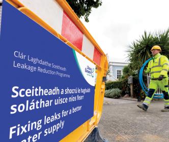 Uisce Éireann carrying a rolled up plastic blue pipe walking across an Uisce Éireann signage about fixing leaks