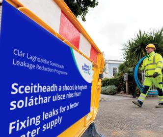 An Uisce Éireann worker carry some blue piping on a site with a sign reading "Fixing leaks for a better water supply"
