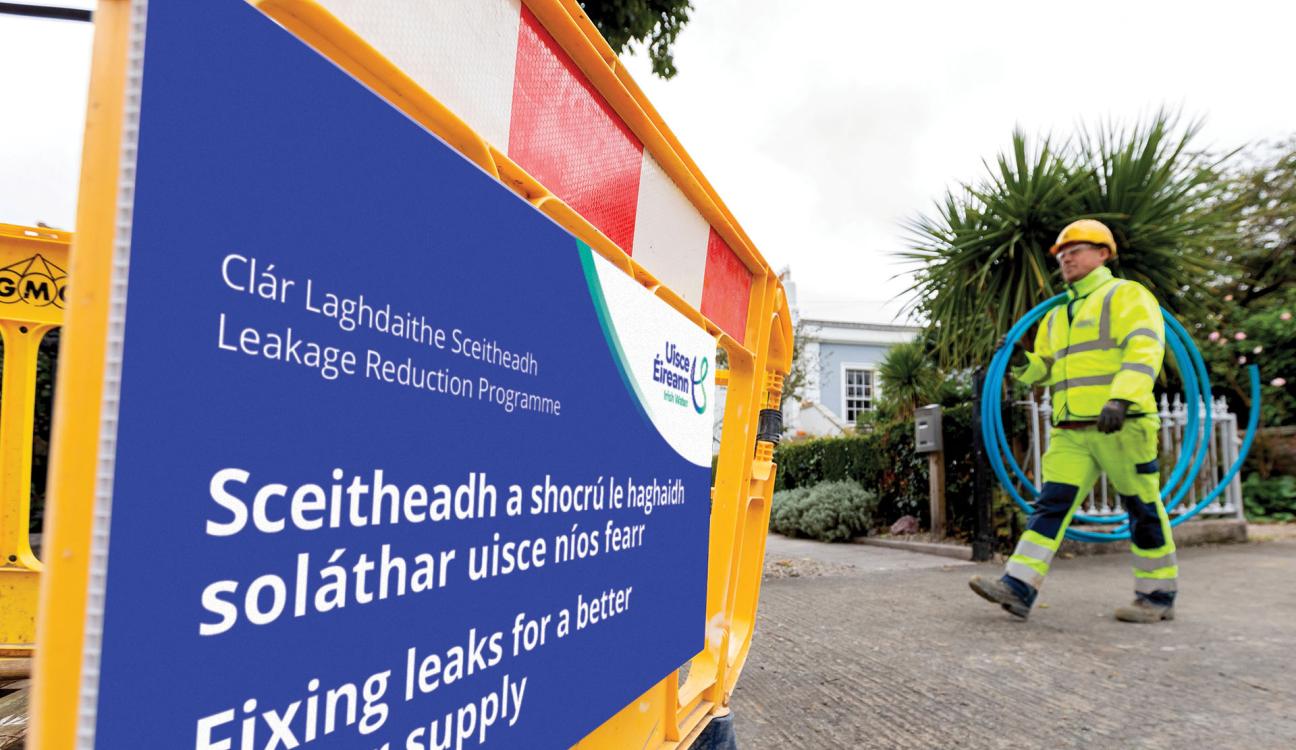 Uisce Éireann carrying a rolled up plastic blue pipe walking across an Uisce Éireann signage about fixing leaks